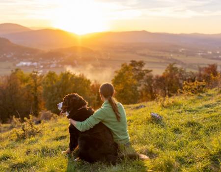 a woman sits beside her dog as they look at the sunset over mountains