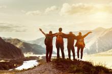 four people, backs turned and arms raised, at the summit of a mountain at sunset